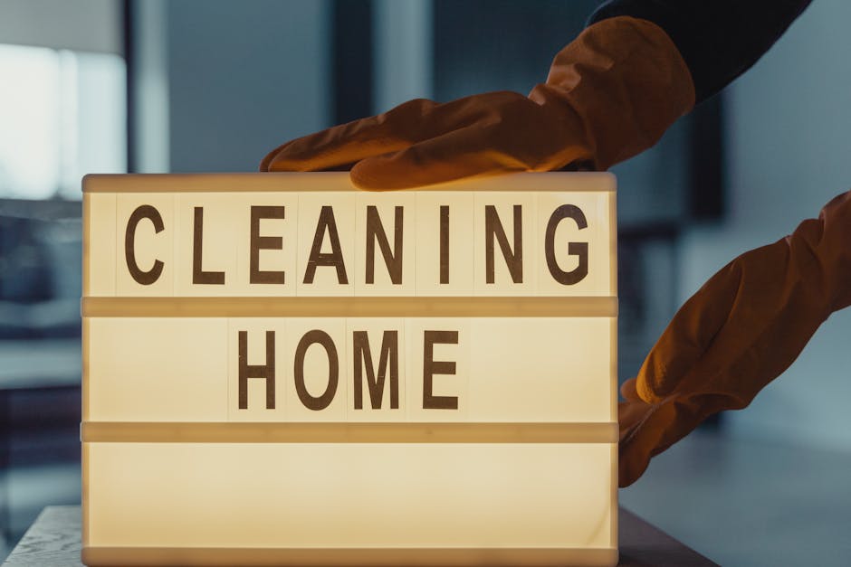 A person wearing yellow rubber gloves is placing a lit, illuminated sign with black letters reading 'CLEANING HOME' on a table, indicating domestic cleaning services. The background shows a blurred in