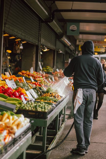 A street-level view of a market stall at Maltby Street market in Bermondsey, focusing on a worker with a dark hoodie and apron spraying a cleaning product onto a display of fresh produce. The stall is