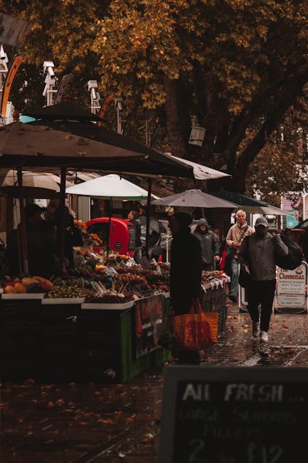 A street-level view of a market stall at Maltby Street market in Bermondsey, focusing on a worker with a dark hoodie and apron spraying a cleaning product onto a display of fresh produce. The stall is