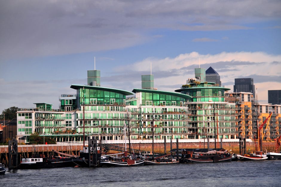 A cityscape view of Surrey Quays featuring modern residential buildings with angular, tiered rooftops and large windows, situated along the water's edge. A tall, cylindrical skyscraper with a glass fa
